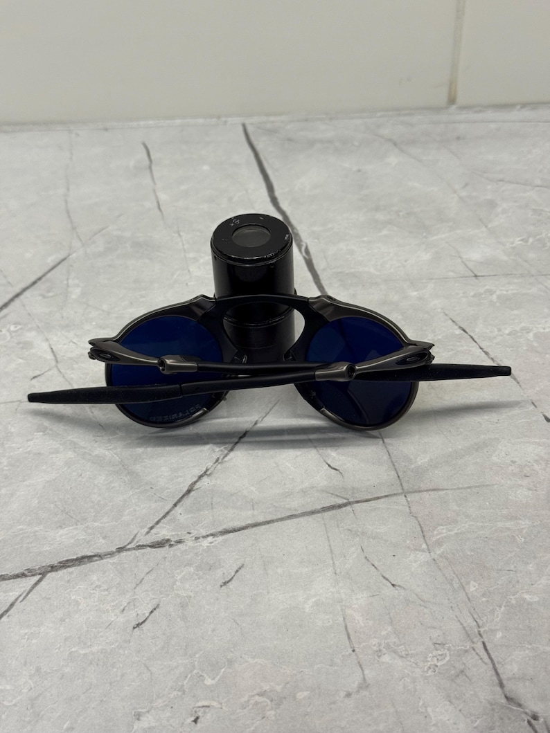 May include: Black sunglasses with blue lenses are displayed on a black cylindrical object. The sunglasses have a sleek, modern design with thin temples and a bridge. The lenses are a vibrant blue color.
