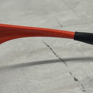 Op de afbeelding: Close-up van de poot van een rode zonnebril met de tekst "MADE IN U.S.A." in het wit gedrukt. De poot heeft een zwarte rubberen punt. De achtergrond is een lichtgrijze ondergrond.