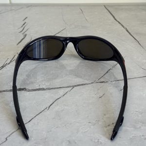 May include: Black sunglasses with dark lenses. The sunglasses have a sleek, sporty design with a curved frame and temples. The lenses are oval-shaped and appear to be tinted. The sunglasses are designed to protect the eyes from the sun.