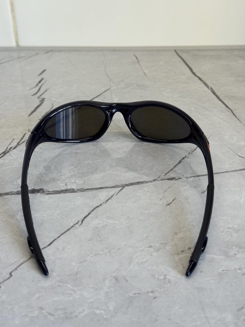 May include: Black sunglasses with dark lenses. The sunglasses have a sleek, sporty design with a curved frame and temples. The lenses are oval-shaped and appear to be tinted. The sunglasses are designed to protect the eyes from the sun.