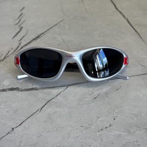 May include: Silver-framed sunglasses with black lenses and red accents on the temples. The sunglasses have a sporty, wraparound design. The brand name "OAKLEY" is visible on the frame.