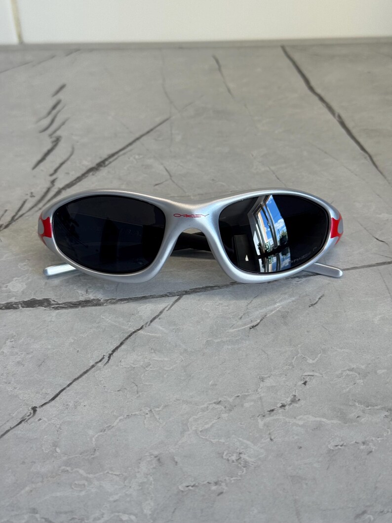 May include: Silver-framed sunglasses with black lenses and red accents on the temples. The sunglasses have a sporty, wraparound design. The brand name "OAKLEY" is visible on the frame.