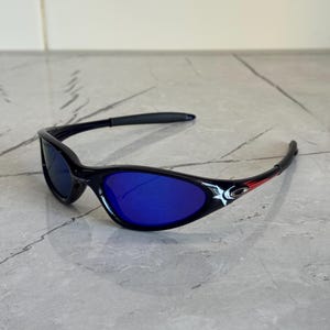 May include: Black sunglasses with blue lenses. The sunglasses have a sleek, curved design with a red accent on the temples. The lenses are a vibrant blue color, and the frames are black.