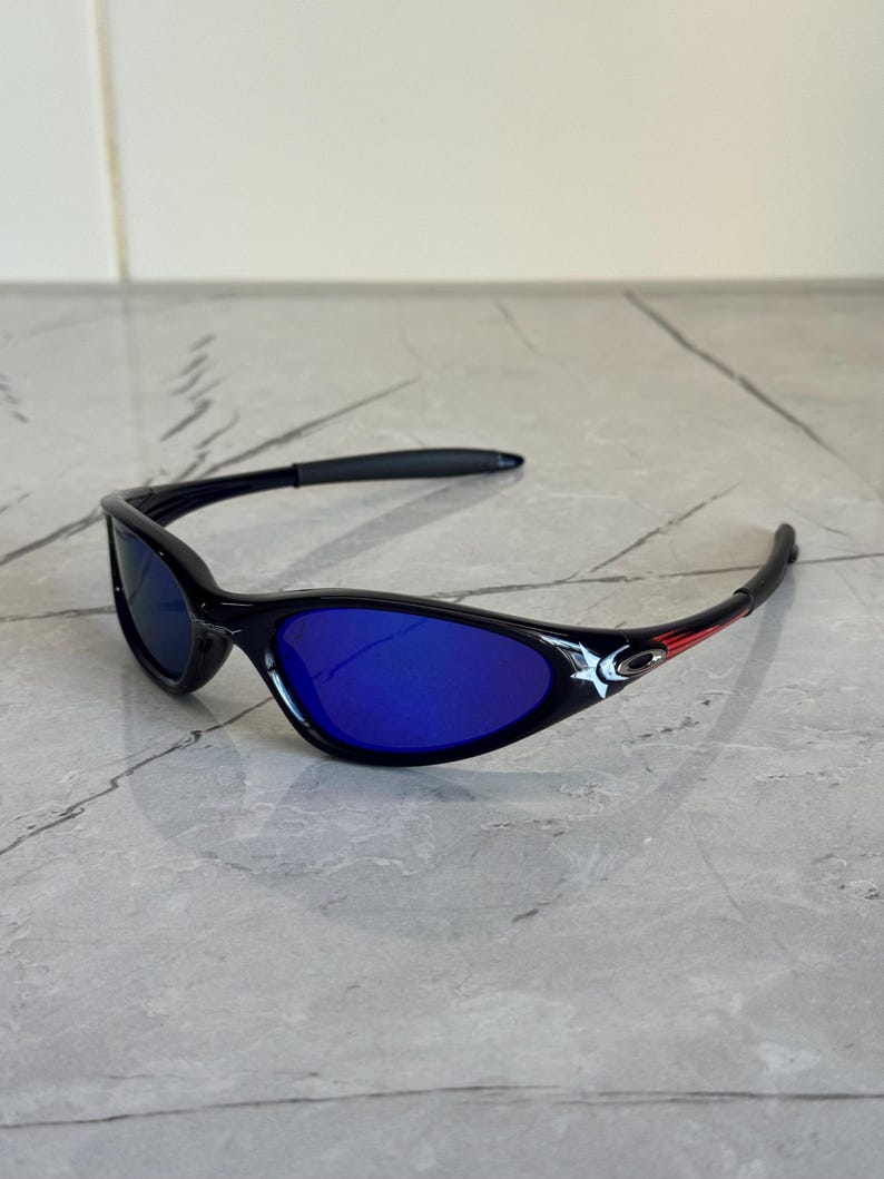 May include: Black sunglasses with blue lenses. The sunglasses have a sleek, curved design with a red accent on the temples. The lenses are a vibrant blue color, and the frames are black.