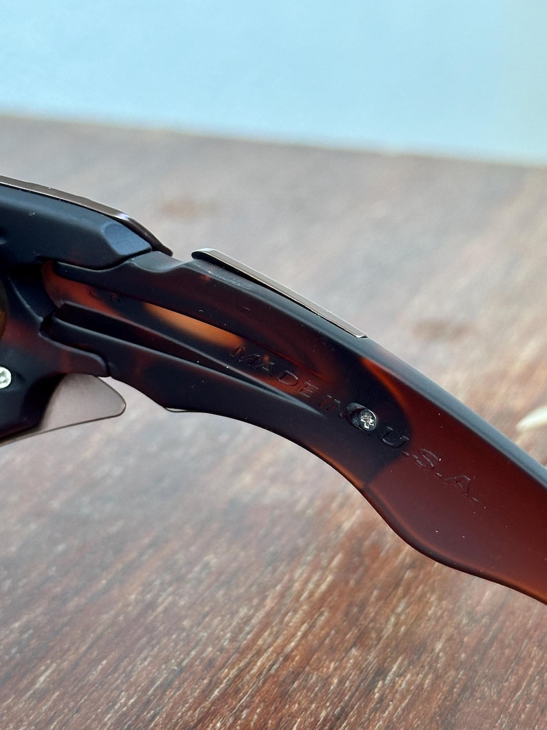Puede incluir: Primer plano de gafas de sol con un patr&oacute;n de carey marr&oacute;n. El texto "MADE IN U.S.A." es visible en la patilla de las gafas. Las gafas de sol est&aacute;n sobre una superficie de madera marr&oacute;n claro.
