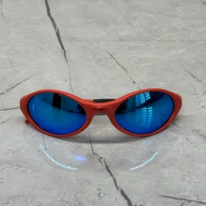 Op de afbeelding: Rode ovale zonnebril met blauwe spiegelglazen. De zonnebril heeft een dik, gebogen montuur en een zwarte brug. De glazen reflecteren de omgeving. De zonnebril ligt op een lichtgrijze ondergrond.