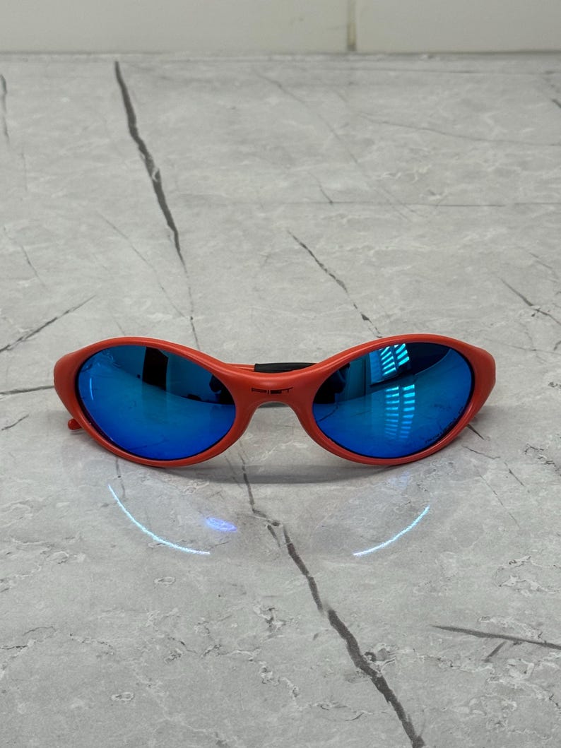 Op de afbeelding: Rode ovale zonnebril met blauwe spiegelglazen. De zonnebril heeft een dik, gebogen montuur en een zwarte brug. De glazen reflecteren de omgeving. De zonnebril ligt op een lichtgrijze ondergrond.