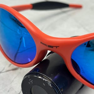 Op de afbeelding: Close-up van een oranje zonnebril met blauwe lenzen. Op het montuur van de zonnebril staat het woord "PIET" gedrukt. De zonnebril rust op een zwart cilindrisch object. De lenzen reflecteren licht.