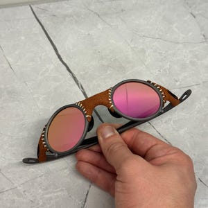 以下が含まれることがあります： 茶色の革のブリッジとテンプルを備えたサングラス。ピンク色のレンズと濃いグレーのフレームが特徴です。革は白い糸で縫われています。サングラスは手で持たれています。