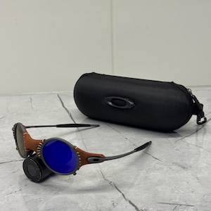 May include: Sunglasses with blue lenses and brown leather accents. The sunglasses have a black frame and are next to a black, oval-shaped case. The case has a zipper and the Oakley logo.