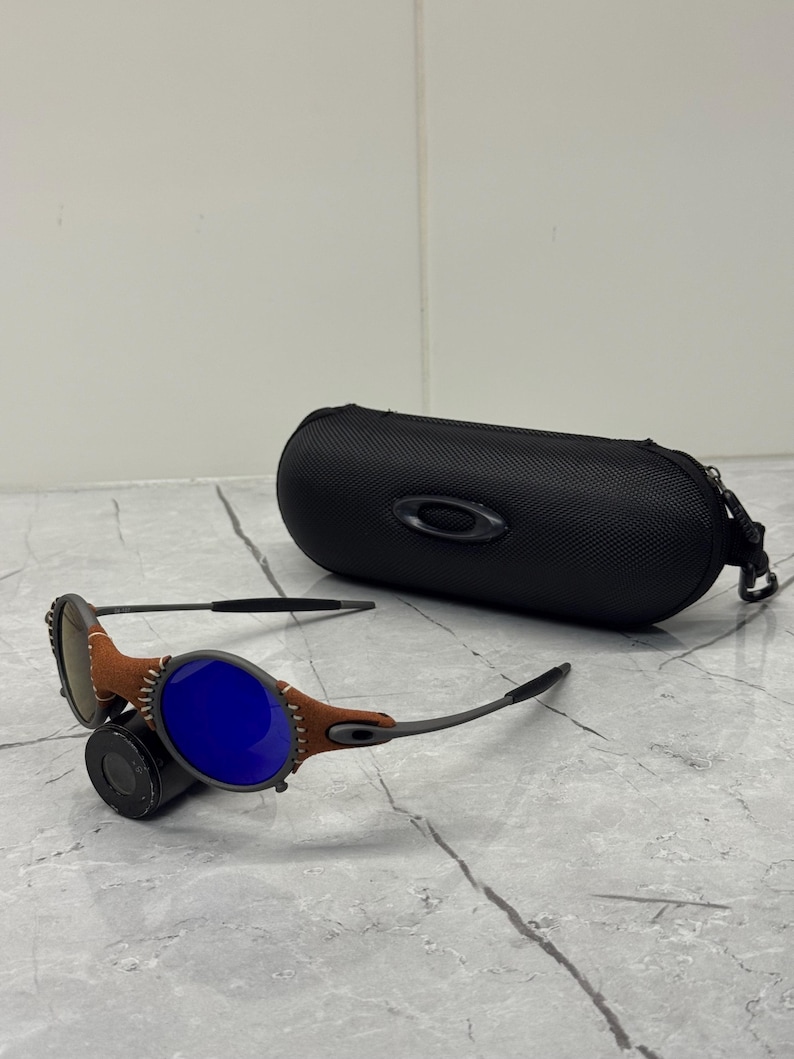 May include: Sunglasses with blue lenses and brown leather accents. The sunglasses have a black frame and are next to a black, oval-shaped case. The case has a zipper and the Oakley logo.