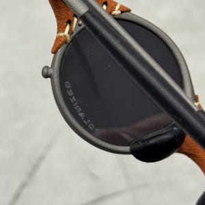 May include: Close-up of sunglasses with a dark gray frame and dark lenses. The temples are covered in brown suede and stitched with cream-colored thread. The lenses have the word "POLARIZED" printed on them.