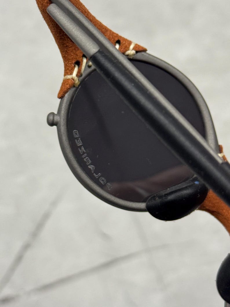 May include: Close-up of sunglasses with a dark gray frame and dark lenses. The temples are covered in brown suede and stitched with cream-colored thread. The lenses have the word "POLARIZED" printed on them.