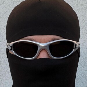 May include: Silver-framed sunglasses with dark lenses. The sunglasses are worn with a black balaclava and a black beanie. The Oakley logo is visible on the frame. The background is a plain white wall.