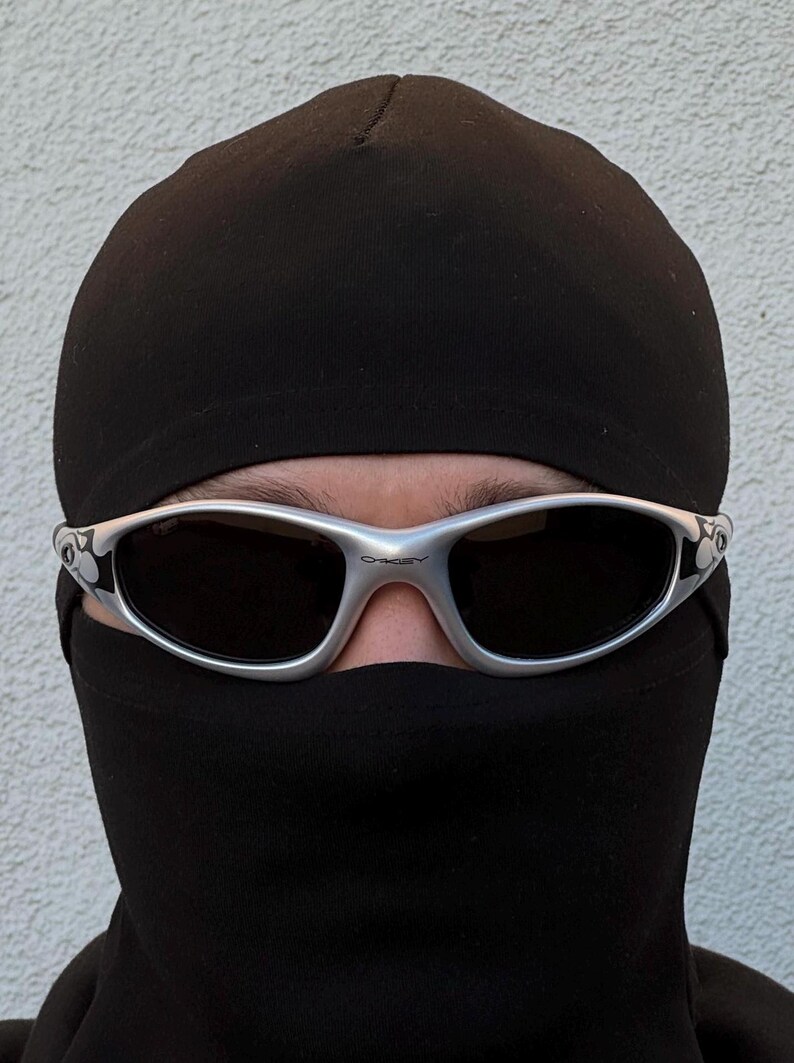 May include: Silver-framed sunglasses with dark lenses. The sunglasses are worn with a black balaclava and a black beanie. The Oakley logo is visible on the frame. The background is a plain white wall.
