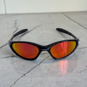 May include: Black sunglasses with a red-orange mirrored lens. The sunglasses have a sleek, sporty design with a black frame and arms. The brand name "Oakley" is visible on the frame.