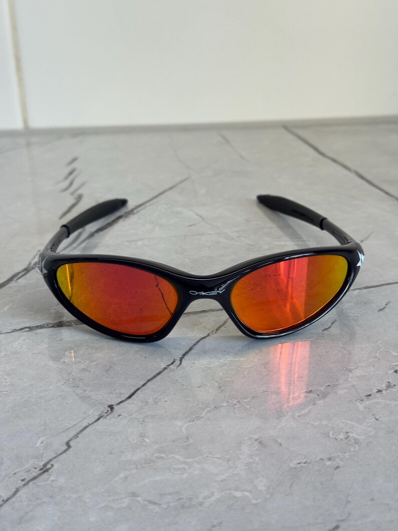 May include: Black sunglasses with a red-orange mirrored lens. The sunglasses have a sleek, sporty design with a black frame and arms. The brand name "Oakley" is visible on the frame.