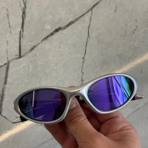 May include: Silver sunglasses with purple lenses. The oval lenses reflect a range of colours. The sunglasses are held in a hand, highlighting the sleek design and modern aesthetic.