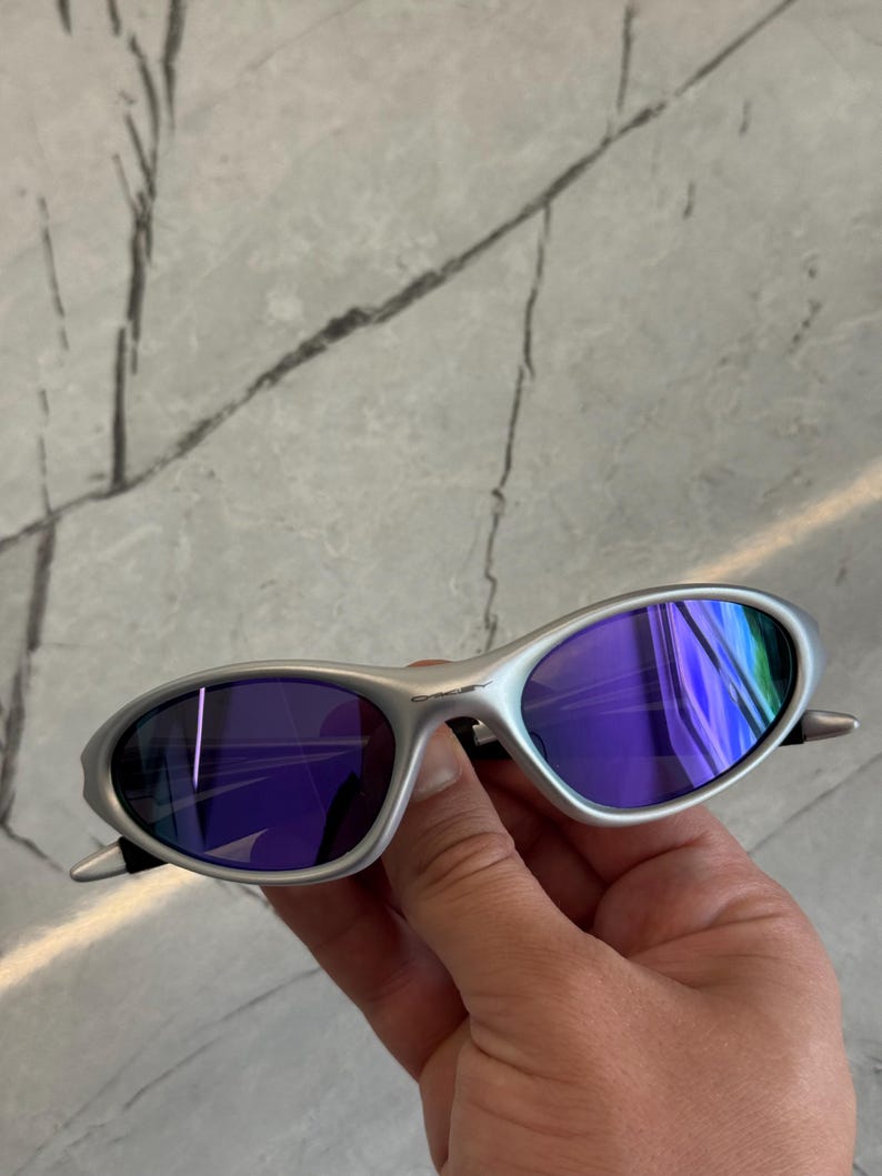 May include: Silver sunglasses with purple lenses. The oval lenses reflect a range of colours. The sunglasses are held in a hand, highlighting the sleek design and modern aesthetic.