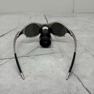 May include: Silver-framed sunglasses with dark lenses. The sunglasses have a sleek, modern design with a curved frame and black temple tips. The sunglasses are resting on a black object.