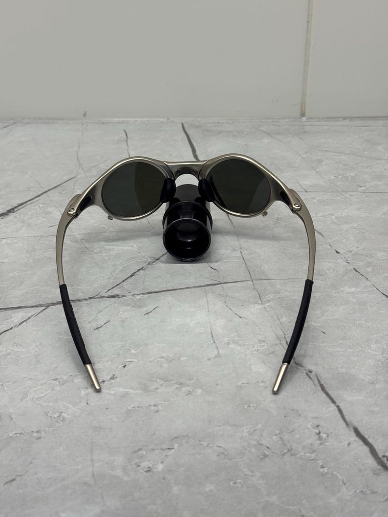 May include: Silver-framed sunglasses with dark lenses. The sunglasses have a sleek, modern design with a curved frame and black temple tips. The sunglasses are resting on a black object.