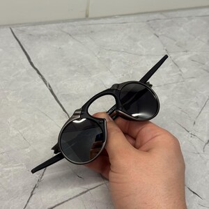 May include: Black sunglasses with a distinctive, angular design. The lenses are round and dark, set in a black frame with silver accents. The arms are thin and extend back from the frame. The sunglasses are held in a hand.