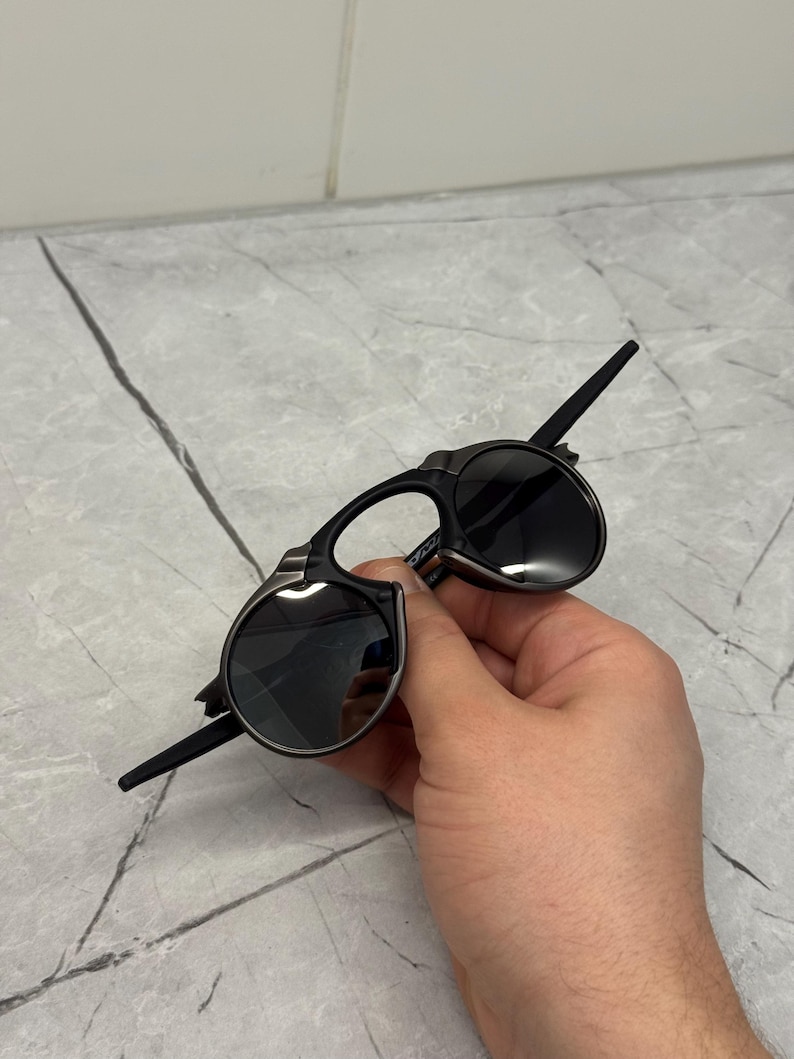May include: Black sunglasses with a distinctive, angular design. The lenses are round and dark, set in a black frame with silver accents. The arms are thin and extend back from the frame. The sunglasses are held in a hand.