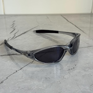 May include: Gray-tinted sunglasses with dark lenses. The sunglasses have a sleek, sporty design with a curved frame and a black nose bridge. The lenses are oval-shaped and provide a contrast to the light gray frame.