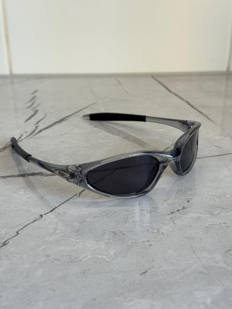 May include: Gray-tinted sunglasses with dark lenses. The sunglasses have a sleek, sporty design with a curved frame and a black nose bridge. The lenses are oval-shaped and provide a contrast to the light gray frame.