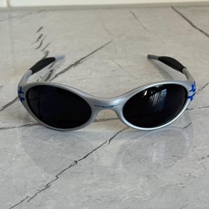 Puede incluir: Gafas de sol plateadas con lentes oscuras. Las gafas tienen un dise&ntilde;o curvo con detalles azules en las patillas y el nombre de la marca impreso en la montura. Las patillas son negras y curvadas para mayor comodidad.