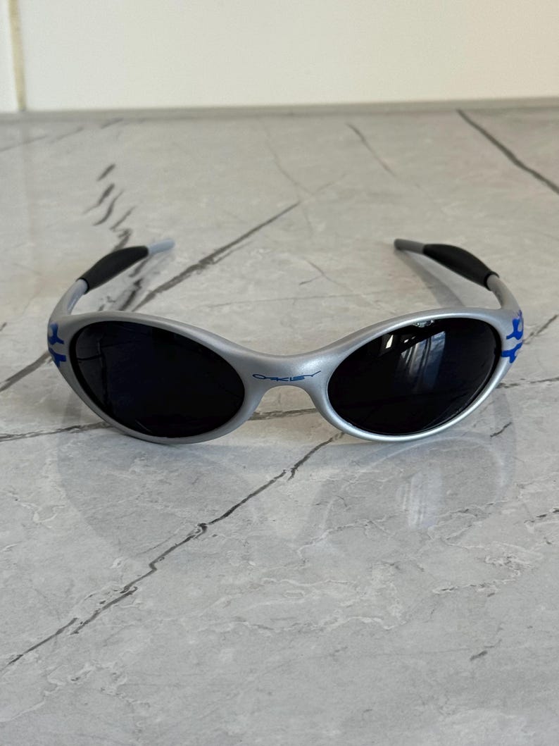 Puede incluir: Gafas de sol plateadas con lentes oscuras. Las gafas tienen un dise&ntilde;o curvo con detalles azules en las patillas y el nombre de la marca impreso en la montura. Las patillas son negras y curvadas para mayor comodidad.