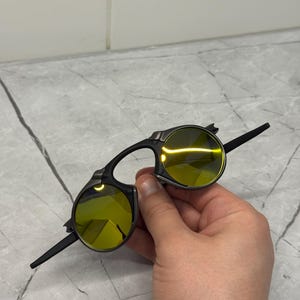 May include: Black sunglasses with yellow-tinted lenses. The sunglasses have a unique design with a bridge and arms that extend from the circular lenses. The lenses are a bright yellow color, and the frames are black.