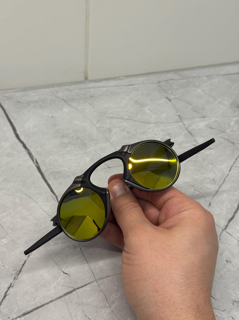 May include: Black sunglasses with yellow-tinted lenses. The sunglasses have a unique design with a bridge and arms that extend from the circular lenses. The lenses are a bright yellow color, and the frames are black.