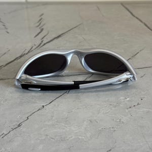 May include: Silver sunglasses with dark lenses. The sunglasses have a sleek, modern design with a curved frame and black accents on the arms. The lenses are dark, providing shade and protection from the sun.