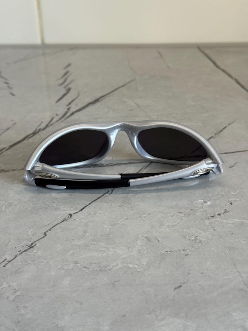 May include: Silver sunglasses with dark lenses. The sunglasses have a sleek, modern design with a curved frame and black accents on the arms. The lenses are dark, providing shade and protection from the sun.