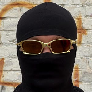 May include: Gold-framed sunglasses with brown lenses are worn over a black balaclava and a black beanie. The background is a weathered brick wall with orange paint.