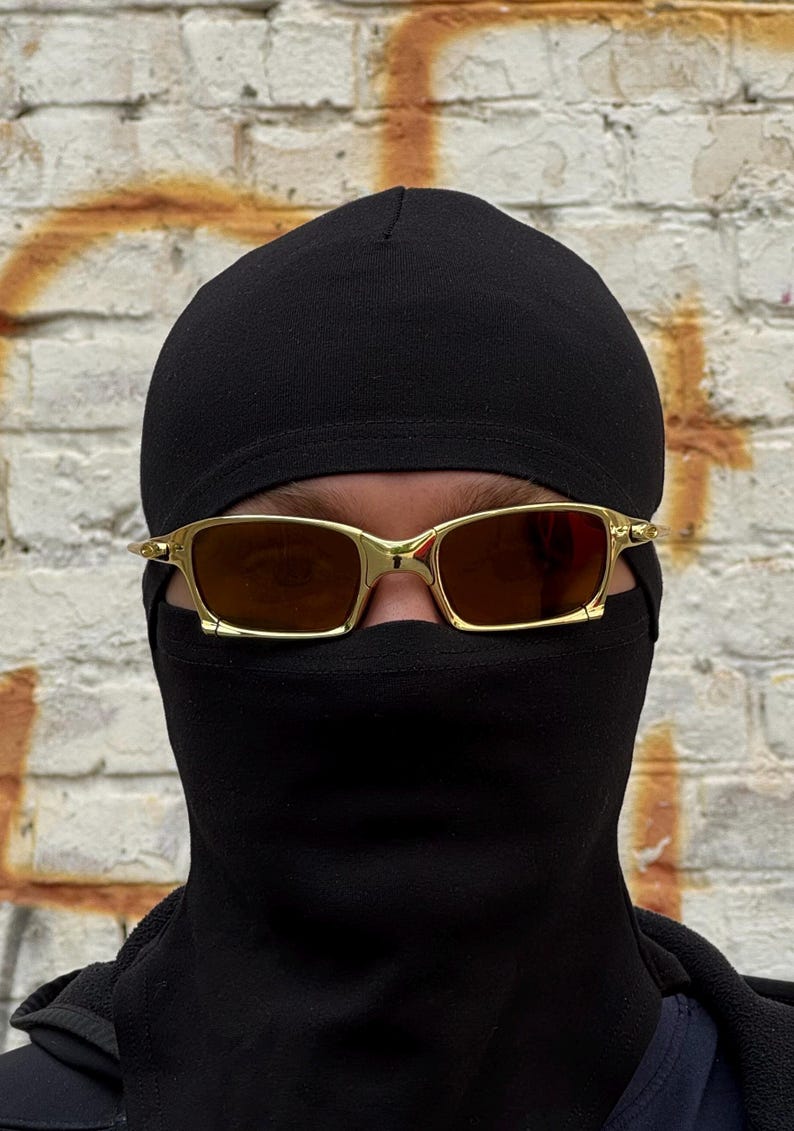 May include: Gold-framed sunglasses with brown lenses are worn over a black balaclava and a black beanie. The background is a weathered brick wall with orange paint.