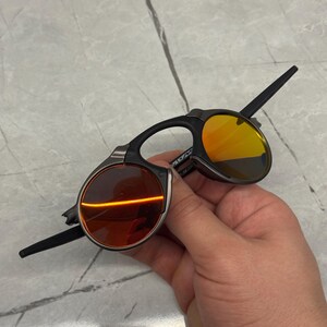 Pu&ograve; includere: Occhiali da sole neri e argentati con lenti arancioni e gialle. Gli occhiali da sole hanno un design unico con una forma di lente rotonda e una montatura elegante e moderna. Le lenti sono di un vibrante colore arancione e giallo.