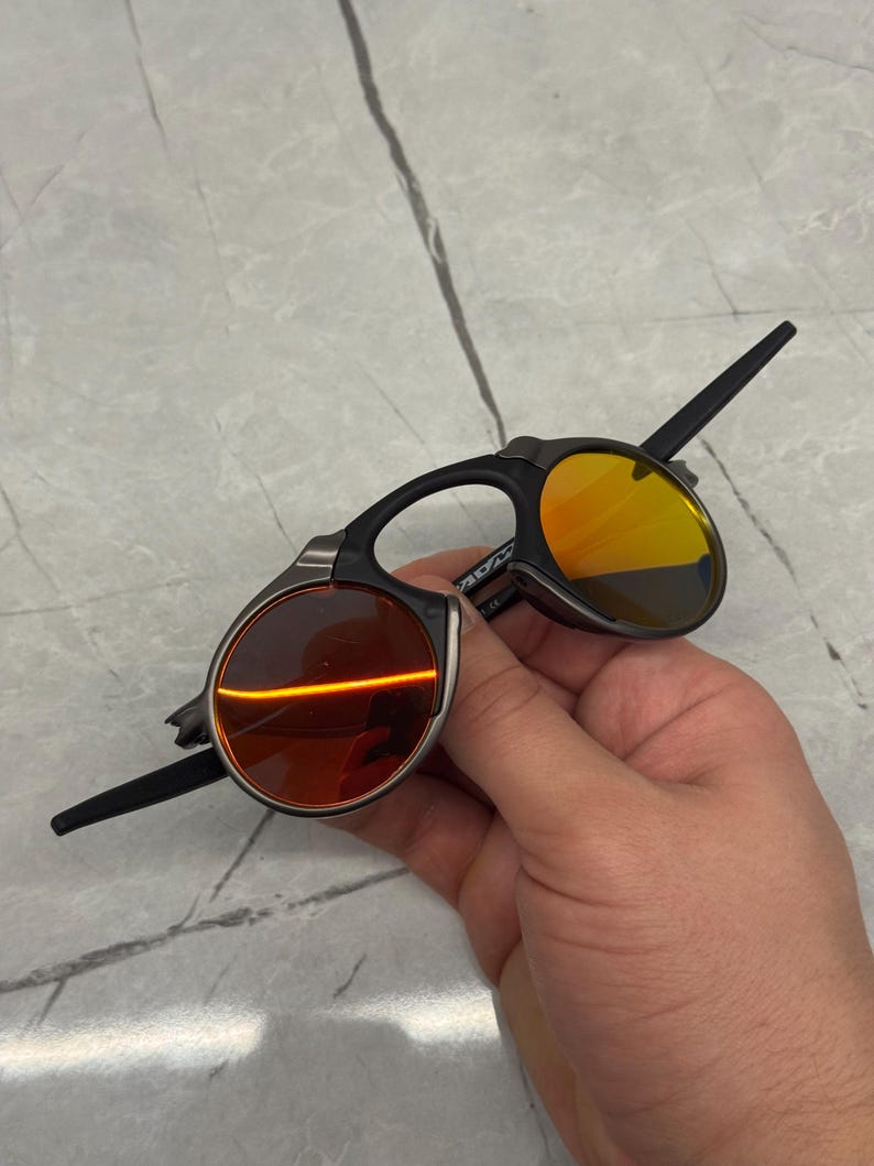Pu&ograve; includere: Occhiali da sole neri e argentati con lenti arancioni e gialle. Gli occhiali da sole hanno un design unico con una forma di lente rotonda e una montatura elegante e moderna. Le lenti sono di un vibrante colore arancione e giallo.