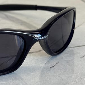 K&ouml;nnte beinhalten: Nahaufnahme einer schwarzen Sonnenbrille mit dunklen Gl&auml;sern. Die Sonnenbrille hat ein schlankes, geschwungenes Design und der Markenname "Oakley" ist auf dem Rahmen sichtbar. Die Sonnenbrille befindet sich vor einem hellen, strukturierten Hintergrund.