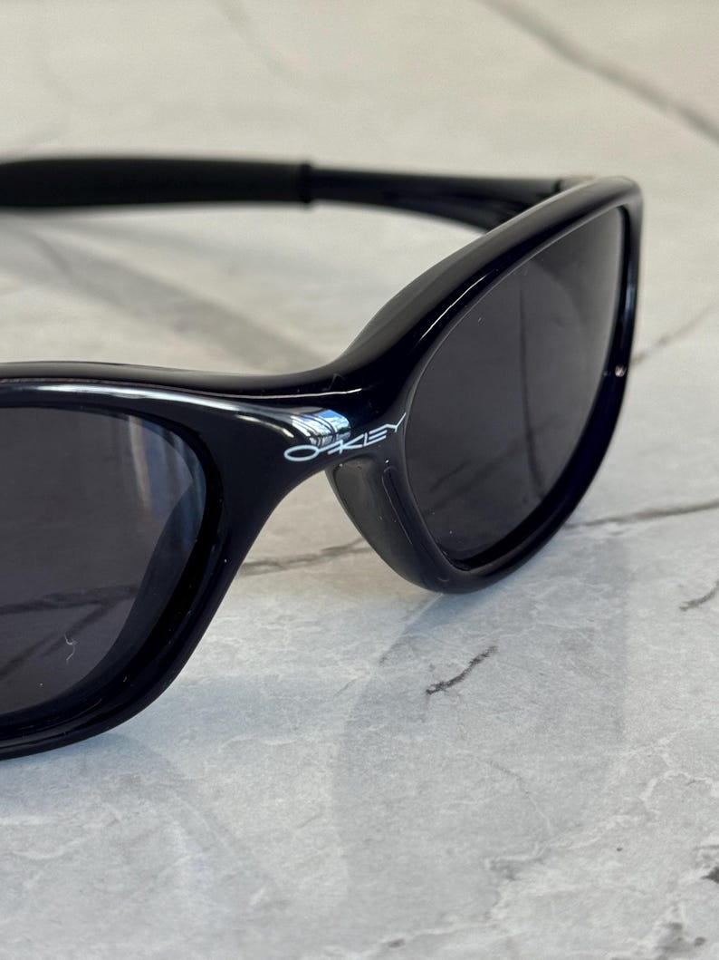 K&ouml;nnte beinhalten: Nahaufnahme einer schwarzen Sonnenbrille mit dunklen Gl&auml;sern. Die Sonnenbrille hat ein schlankes, geschwungenes Design und der Markenname "Oakley" ist auf dem Rahmen sichtbar. Die Sonnenbrille befindet sich vor einem hellen, strukturierten Hintergrund.