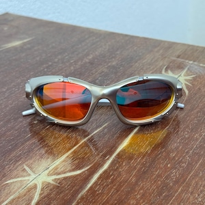 Puede incluir: Gafas de sol con montura plateada y lentes naranjas. Las gafas tienen un dise&ntilde;o futurista &uacute;nico con una montura curvada y patillas blancas peque&ntilde;as. Las lentes reflejan el entorno, mostrando una mezcla de tonos naranjas y azules.