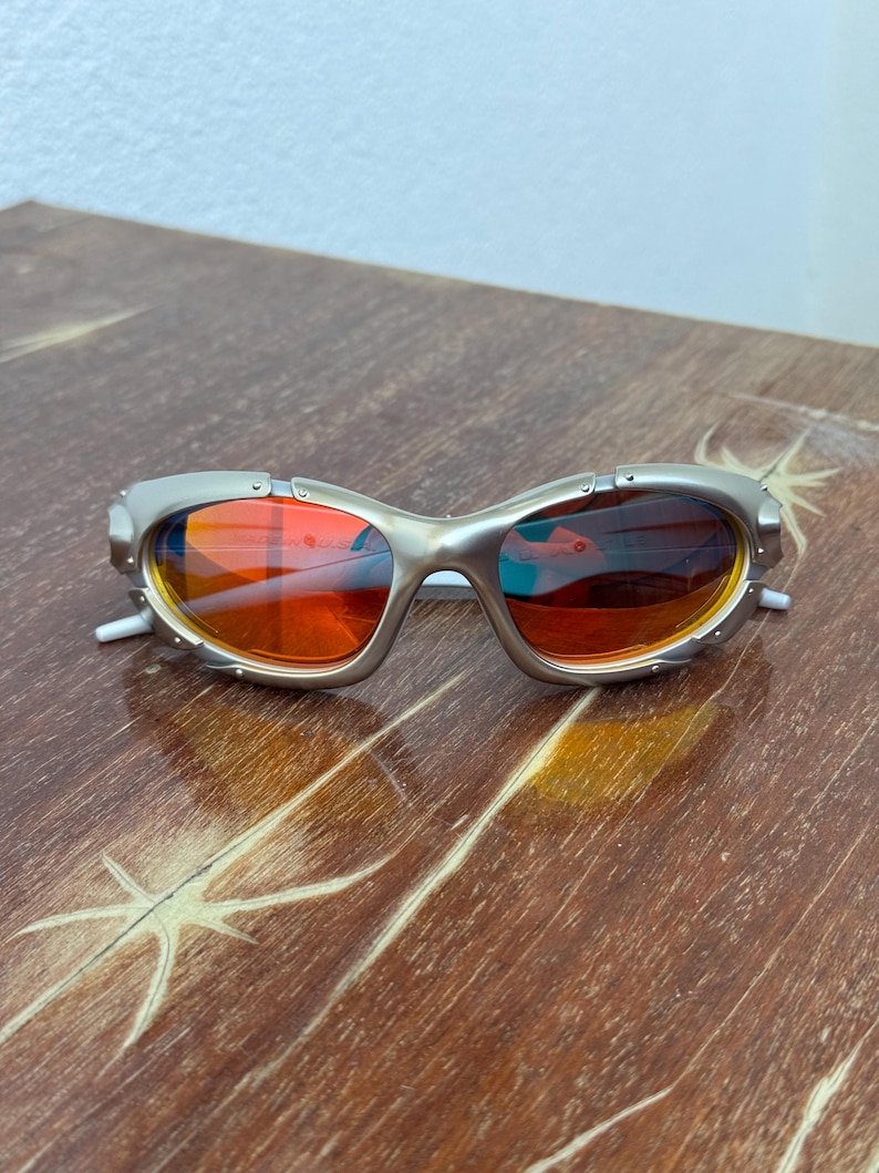 Puede incluir: Gafas de sol con montura plateada y lentes naranjas. Las gafas tienen un dise&ntilde;o futurista &uacute;nico con una montura curvada y patillas blancas peque&ntilde;as. Las lentes reflejan el entorno, mostrando una mezcla de tonos naranjas y azules.