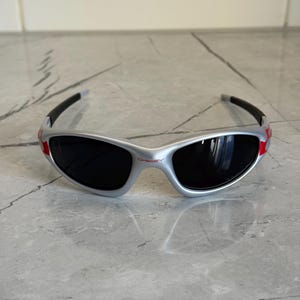 May include: Silver-framed sunglasses with black lenses and red accents. The sporty eyewear features a wraparound design and a subtle logo on the bridge. The temples are black and curve around the ears.