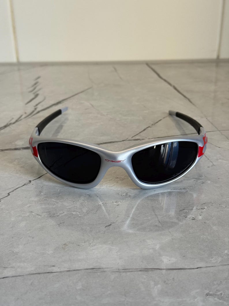 May include: Silver-framed sunglasses with black lenses and red accents. The sporty eyewear features a wraparound design and a subtle logo on the bridge. The temples are black and curve around the ears.