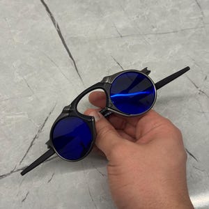 以下が含まれることがあります： 青いレンズの黒とシルバーのサングラス。 サングラスはユニークで角張ったフレームデザインです。 レンズは丸く、光を反射します。 手で持っています。