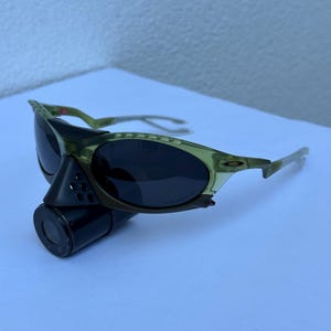 May include: Olive green sunglasses with dark lenses. The sunglasses have a sporty design with a curved frame and a black cylindrical attachment. The lenses are oval-shaped.