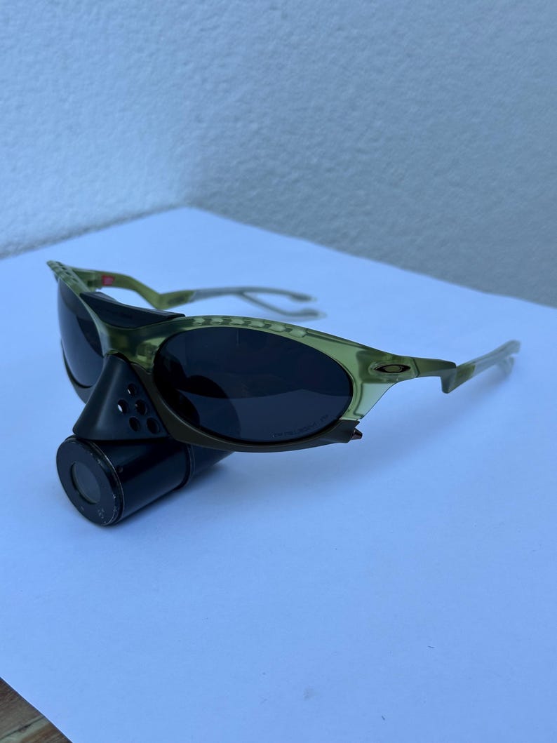 May include: Olive green sunglasses with dark lenses. The sunglasses have a sporty design with a curved frame and a black cylindrical attachment. The lenses are oval-shaped.