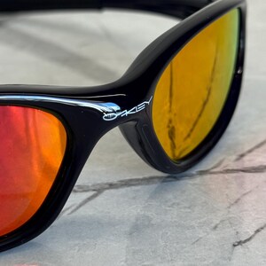 May include: Close-up of black sunglasses with orange and yellow gradient lenses. The brand name "Oakley" is visible on the frame. The sunglasses have a sleek, modern design.