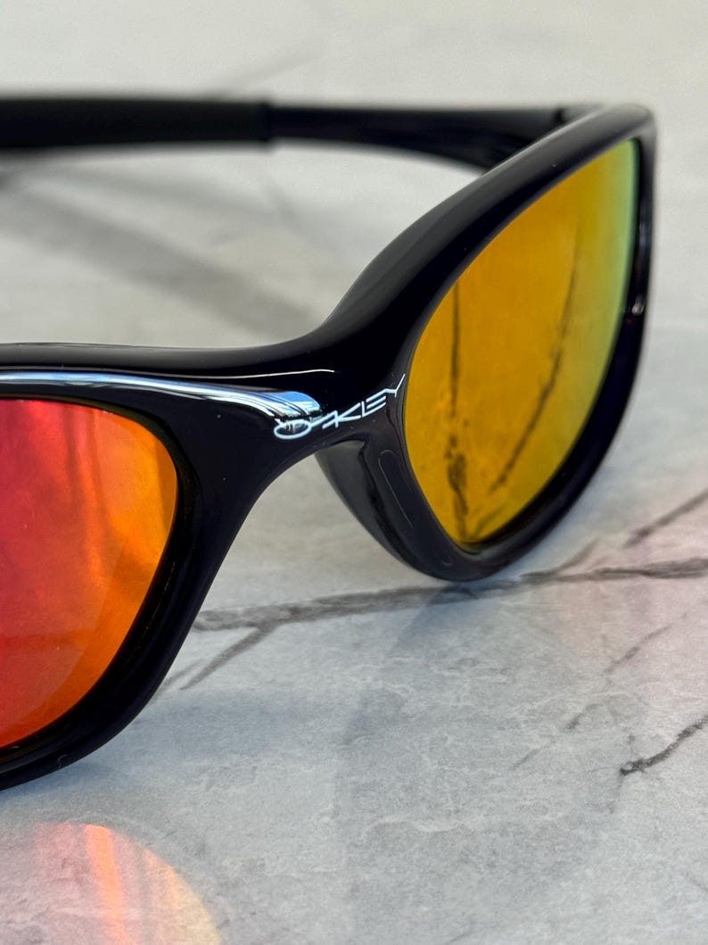 May include: Close-up of black sunglasses with orange and yellow gradient lenses. The brand name "Oakley" is visible on the frame. The sunglasses have a sleek, modern design.